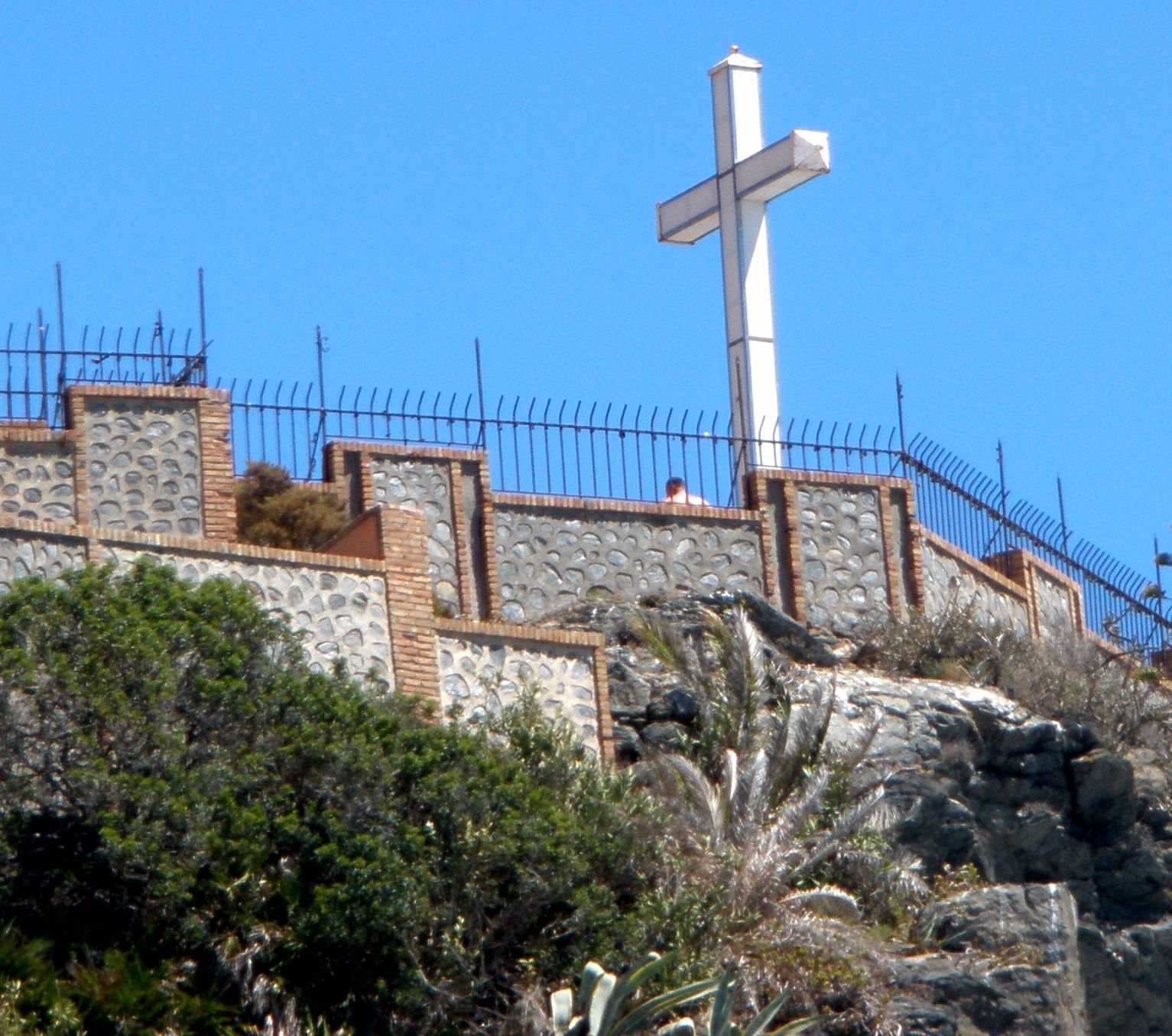 Korset på Peñón del Santo, højt over murene på udsigtspladsen. Se godt efter på den skrå stang i gelænderet ude til højre: den er tyk af hængelåse